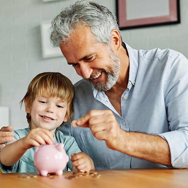 Grandparent And His Grandson Putting Coin Money In Piggybank At Home. Personal Savings, Bank Safety And Financial Investments Concept